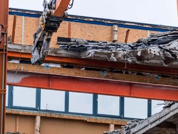 Construction machinery demolishing a building with exposed concrete and steel beams.