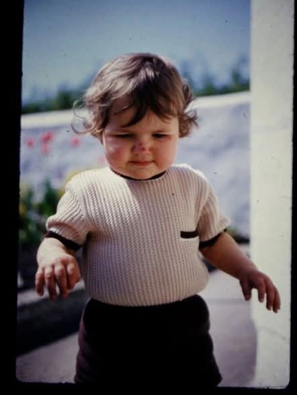 Toddler with curly hair wearing a knitted sweater outdoors.