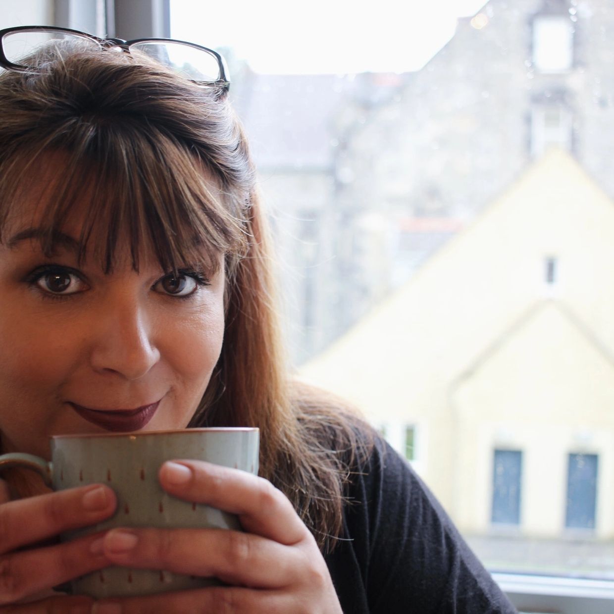 Woman enjoying a warm drink by the window with glasses on her head.