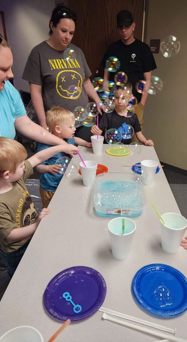 Children and adults play with bubbles at a table indoors.