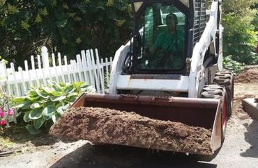 A small Bobcat loader carrying soil near a white picket fence.