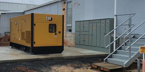 Industrial generator and electrical panels outside a building under cloudy sky.