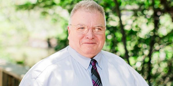 Smiling man in glasses wearing a white shirt and patterned tie outdoors.