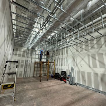 Two workers installing metal framework on a ceiling in a large unfinished room.