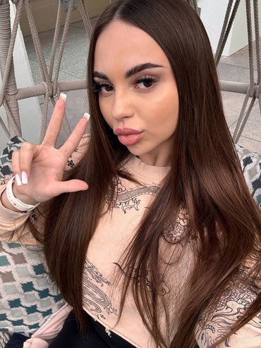 Woman with long brown hair making a peace sign while sitting on a hanging chair.