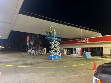 Workers repair the gas station canopy at night using a scissor lift.