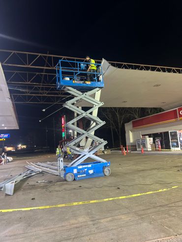 Worker on a blue scissor lift at a gas station under nighttime construction.