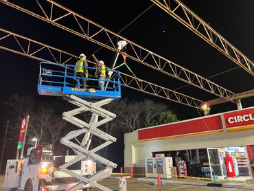 Workers on a scissor lift and rooftop welding at a gas station at night.