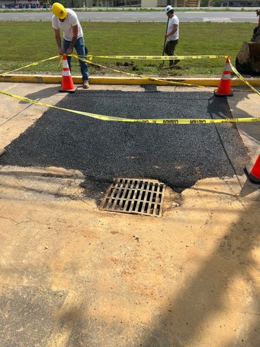 Workers repairing a section of road near a drainage grate with caution tape and cones.