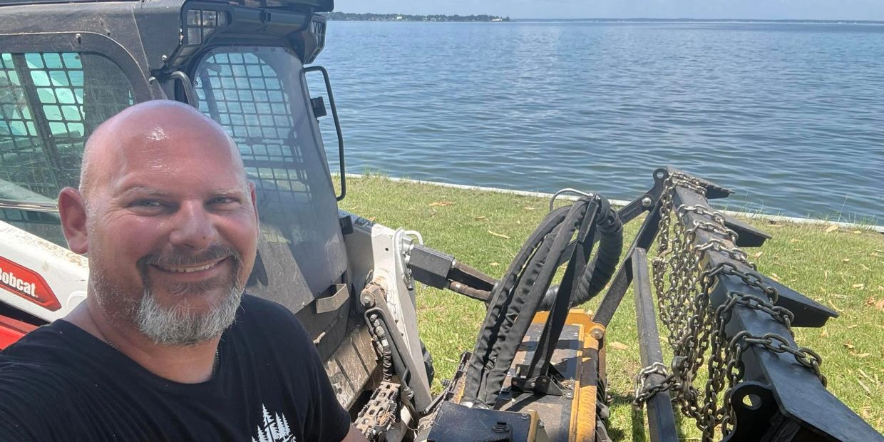 Man smiling beside a Bobcat machine near water.