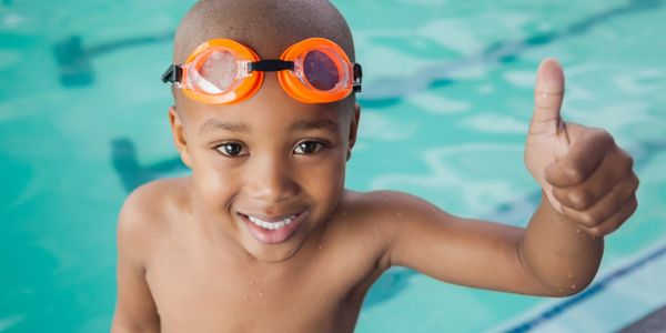 Young boy in a pool with goggles, smiling and giving a thumbs up.