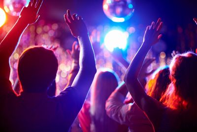 People at a dance party holding their hands in the air while dancing under lights and a disco ball.