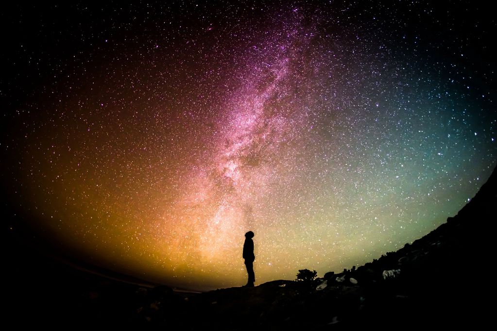 A person stands under a vast, multicolored night sky filled with countless stars.