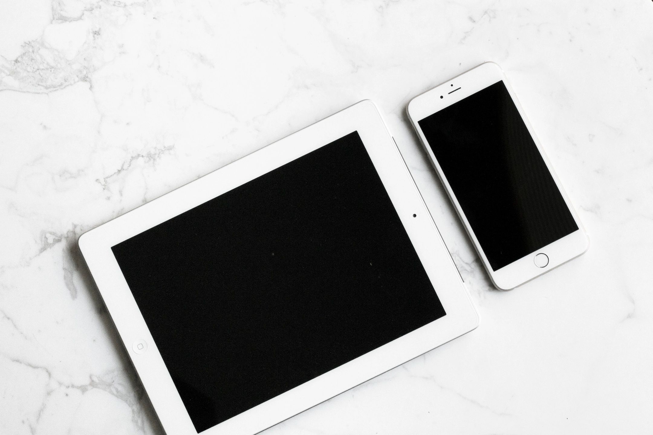 White colored tablet and smartphone sitting side by side on a marble counter top.