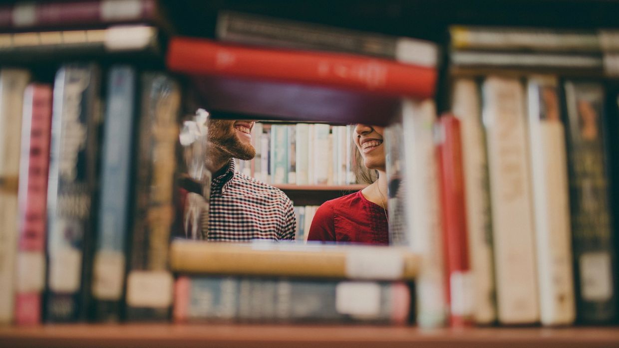 two people seen smiling at each other through a stack of books