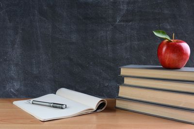 a stack of books with an apple on top next to a blank notebook,open with a pen on top
