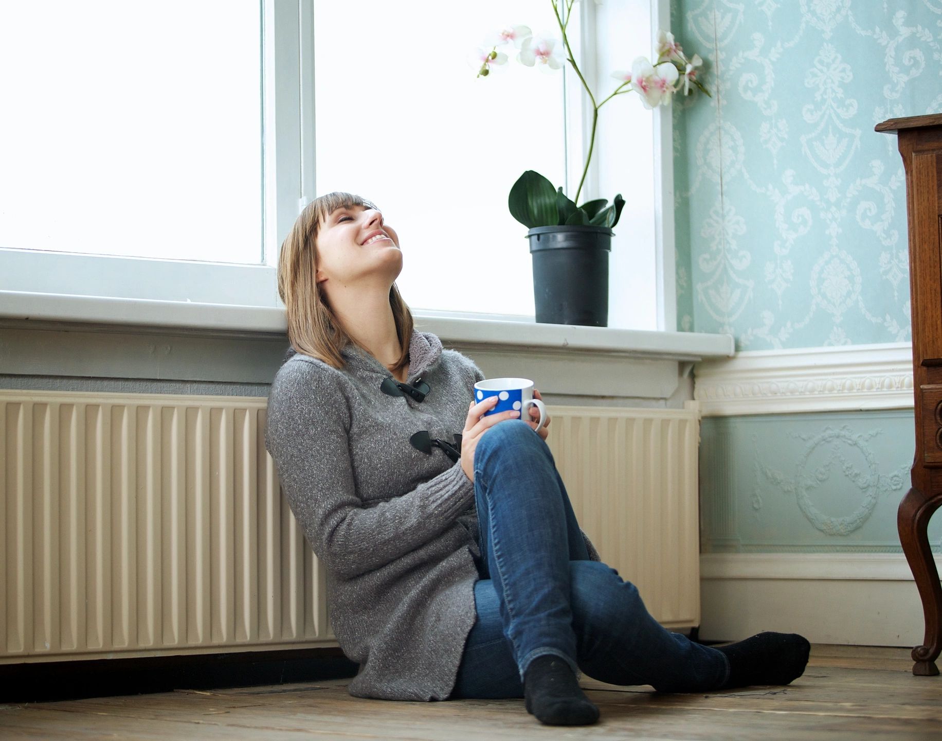 woman relaxed and smiling, sitting on the floor, against the radiator, flower pot on window sill 