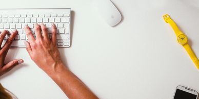 A person typing on a keyboard with an apple mouse, yellow watch and cell phone.