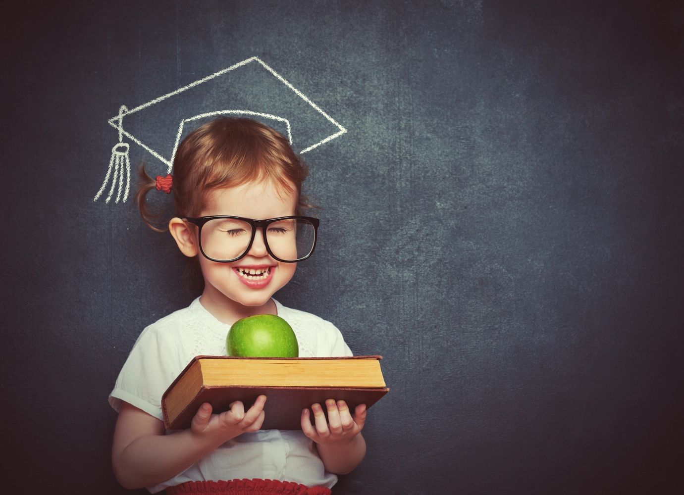 Child smiling wearing big framed glasses holding apple & book in front a of chalk graduation hat
