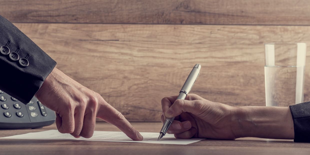 Photo of hands- one signing a document, the other pointing where to sign.