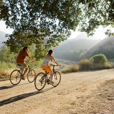 A women and a man, each riding a bicycle on dirt road in the country with a tranquil setting.