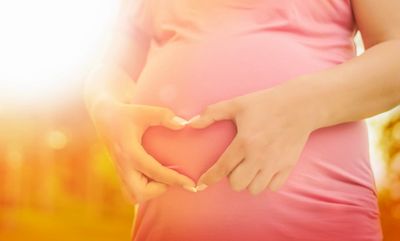 A woman is holding her fingers in a heart shape in front of her pregnant abdomen; radiant sunlight.