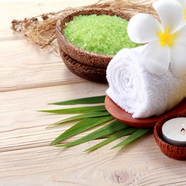 A table with sugar scrub, a towel, candle and tincture decorated with white flowers and palm fronds