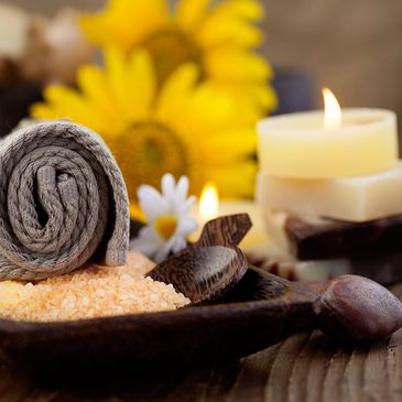 Gray spa towel surrounded by bath salts, yellow and white flowers, soaps topped with a burning candl