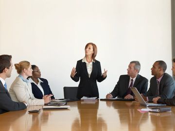 Group of seven person sitting at table in meeting setting with computers and paperwork 