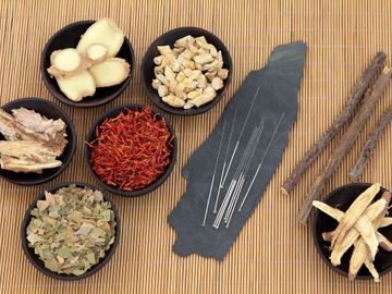Small bowls of chinese herbs, acupuncture needles on a flat stone, all on a reed mat