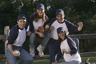 Group of four people sitting on fence in baseball shirts holding a baseball bat, softball and glove.
