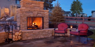 Outdoor fireplace built from stone with a fire glowing across 2 chairs on the natural stone patio