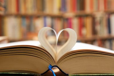 Photo of an open book in front of bookshelves, with two pages curled to make a heart shape.