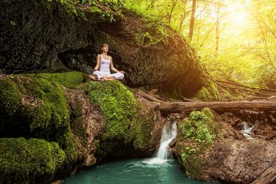 Una mujer en posición de meditación sentada delante de un río en un bosque verde con el sol detrás.