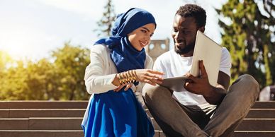 A young person wearing a blue hijab and white jacket helps a young Black man with a laptop. 