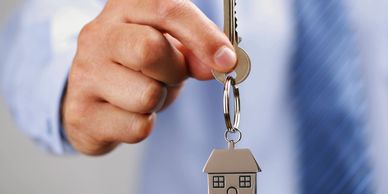 a man in a dress shirt extends a key on a keychain in the shape of a house toward the viewer