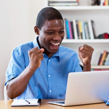 A man holding up two fists in excitement while looking at his laptop on the table, notebook and pen laying to the right.