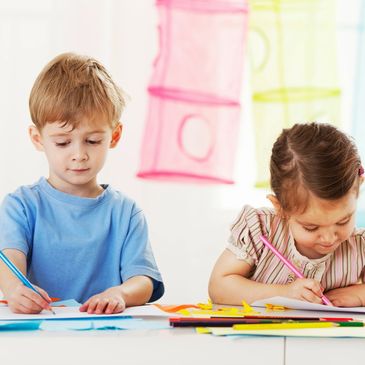 A boy is coloring with a crayon and a girl is writing on the paper with a pencil.