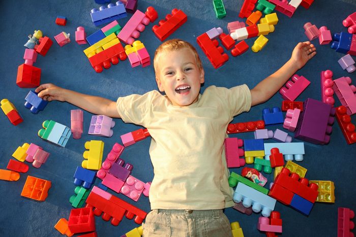 Boy laying on ground with large colourful Lego bricks surrounding him. His a expression is happiness