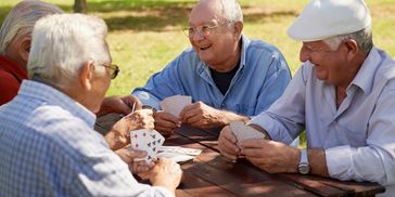 Group of older men sitting at picnic table on nice weather day playing cards and laughing