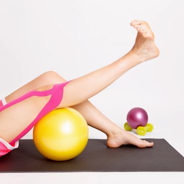 A leg with pink kinesiology tape extends over a ball on a mat, with weights nearby.