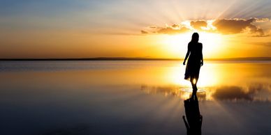 The back side of a woman walking. Background of reflective sunset with hues of yellow and blue sky.