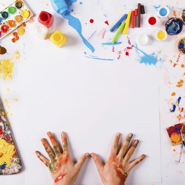 A blank white large piece of art paper surrounded by messing paints with two hands on top 