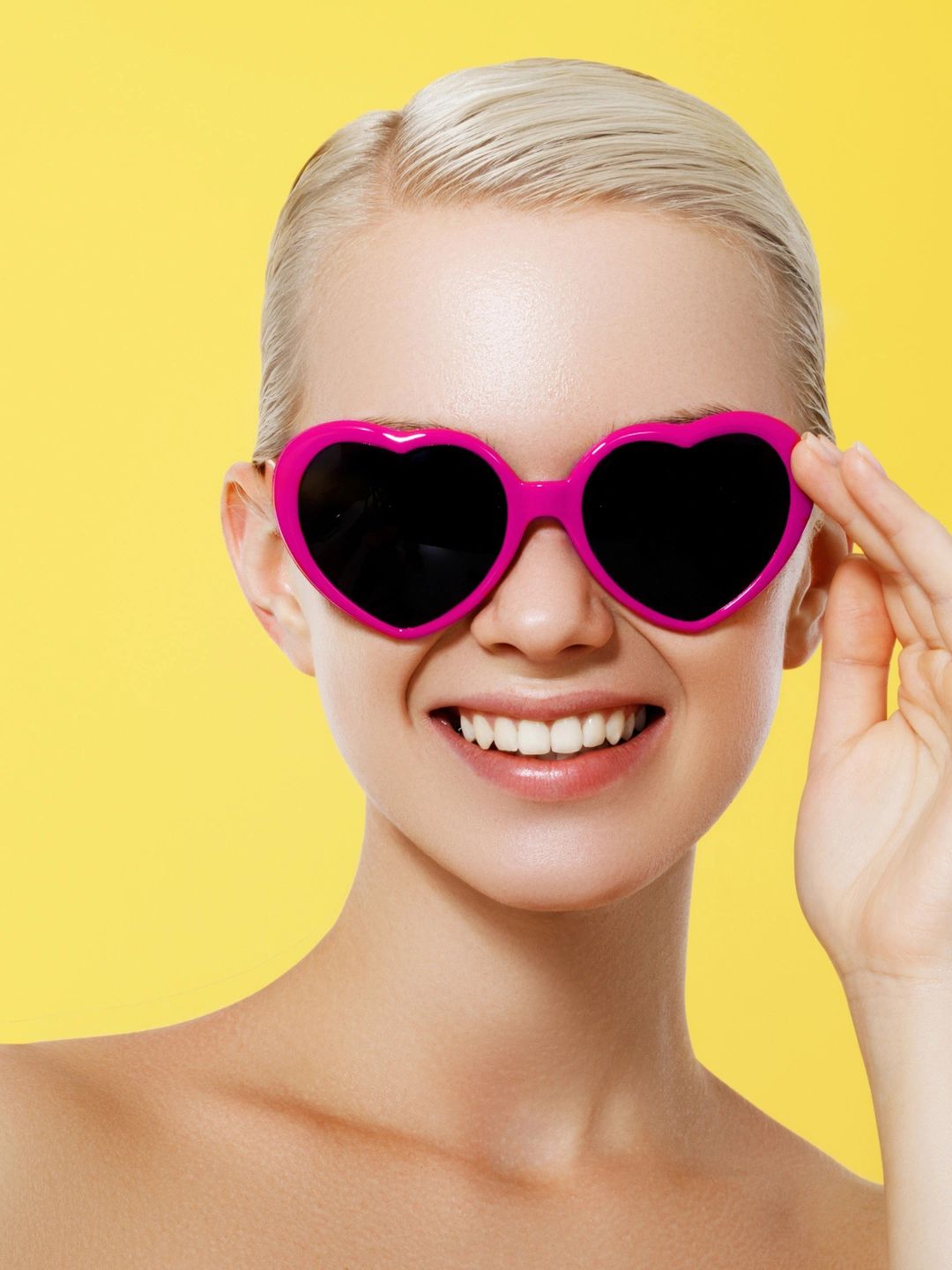 A woman with short blonde hair smiling on a yellow background. Wearing heart shaped pink sunglasses.