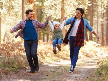Two men walk in a forested area, smiling and swinging a young child between them.