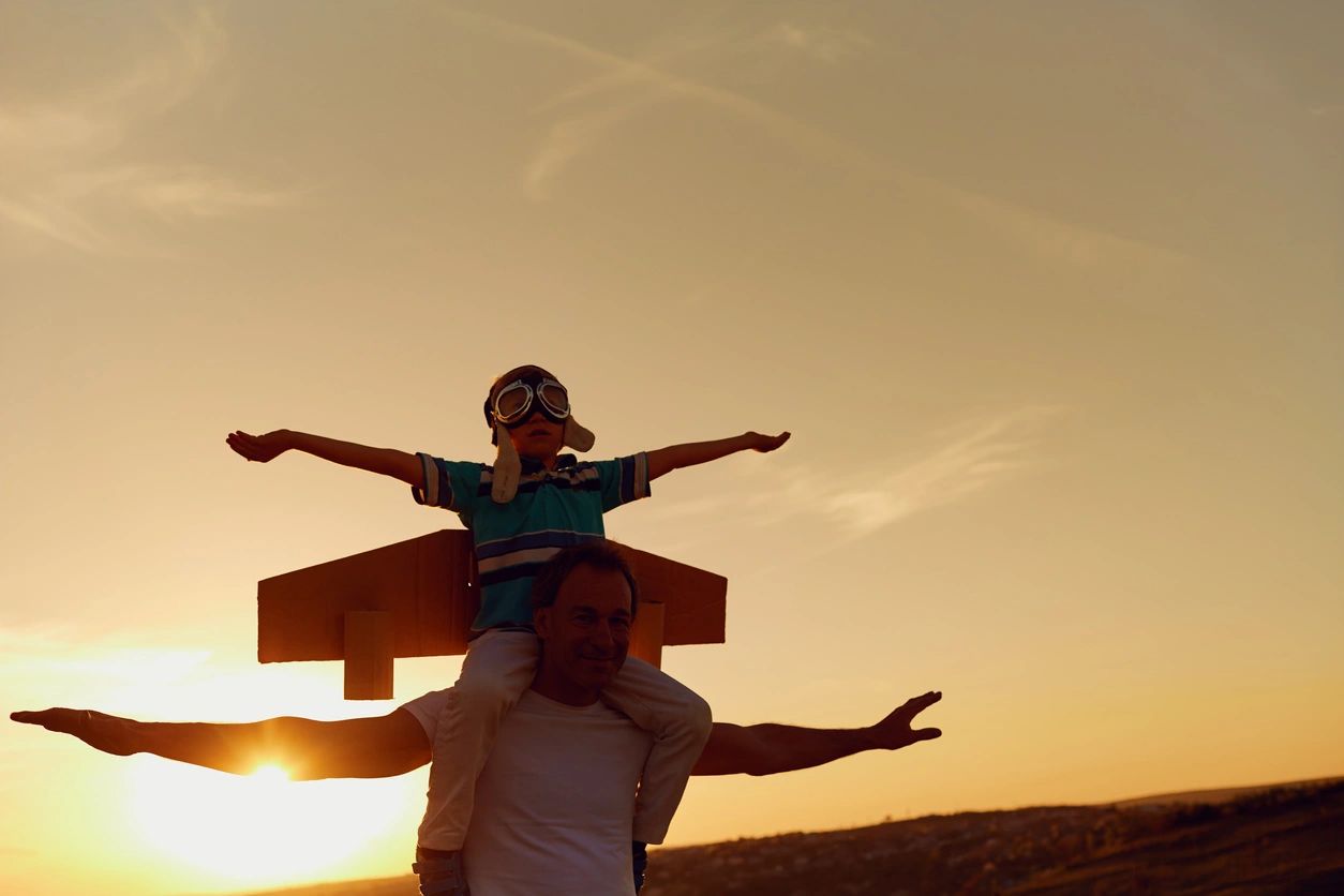 Father with son riding on his shoulders, both with arms outstretched, in blazing sun, low in the sky