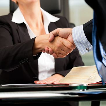 A female attorney sitting at desk with law book and shaking the hand of a male attorney standing. 
