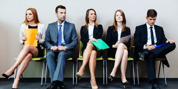 Three women and two men sitting in chairs against a wall waiting for a job interview.
