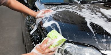 A person is washing with a soapy foam hand brush a black car.