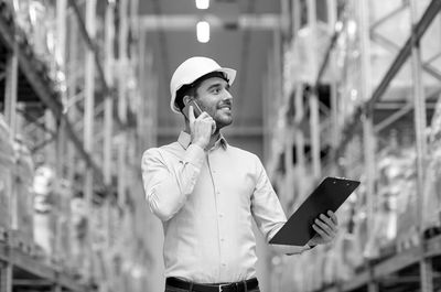 Project Manager holding clipboard whilst on phone, wearing a hardhat in a warehouse.